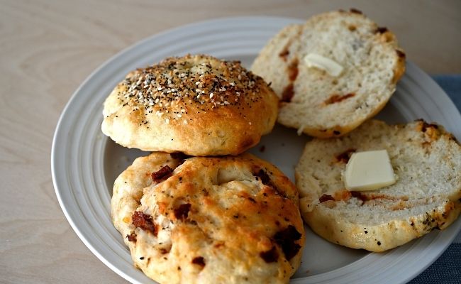 A sundried tomato and basil bagel cut in half with butter on a plate with two other bagels.