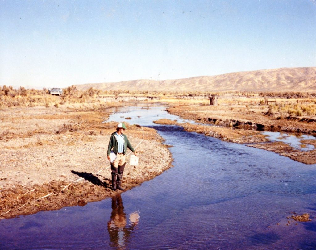 A man standing near a small river in an arid dessert and mountainous landscape.